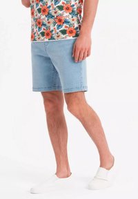 Shorts en denim bleu clair à coupe droite, avec un ourlet brut et des poches latérales, associés à une chemise à imprimé floral blanc et des chaussures à enfiler blanches.