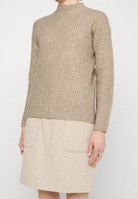 Pull beige en maille avec un col montant, une texture côtelée, des manches longues et une coupe décontractée, associé à une jupe trapèze beige clair avec des poches.