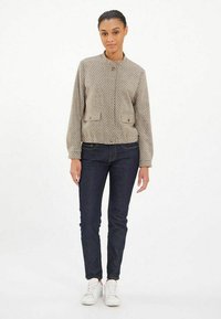 Veste bomber à motifs beige avec un col arrondi, deux poches avant et fermeture à boutons, assortie à un jean bleu foncé et des baskets blanches.