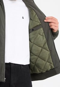 Personne portant un pantalon noir et une chemise blanche ouvre une veste matelassée vert olive, montrant la doublure rembourrée et la poche pression à l’intérieur.