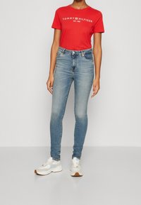 Camiseta roja de Tommy Hilfiger con logo blanco, combinada con jeans ajustados de color azul claro y zapatillas blancas con detalles marrones, de pie contra un fondo sencillo.