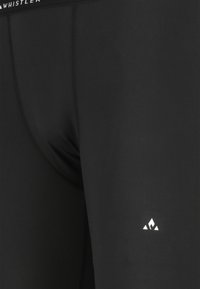 Tæt på sorte atletiske leggings med synlige sømme og et lille hvidt trekantet logo på venstre lår nær taljen mærket "WHISTLER."
