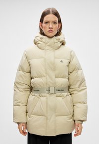 Beige pufferjacka med hög krage, bälte i midjan och stora framfickor. Har en slät, matt textur och subtil logotyp.