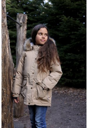 Bež zimska parka z ovratnikom obloženim z umetnim krznom, sprednjimi gumbi in stranskimi žepi. Nošena preko kavbojk na gozdnem ozadju.