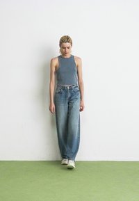 Jeune femme avec des cheveux blonds portant un haut sans manches gris, un jean bleu ample et des baskets blanches marchant devant un mur blanc.