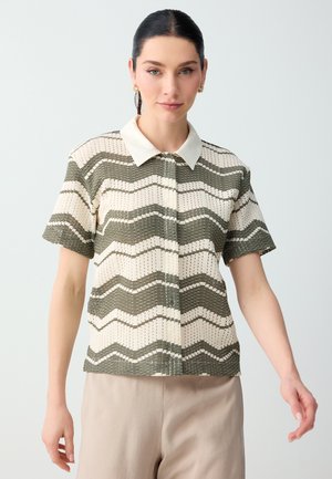 Vrouw draagt een kort mouwens, gebreide top met kraag met olijfgroene en crèmekleurige golvende strepen, gecombineerd met lichtbeige broek tegen een effen achtergrond.
