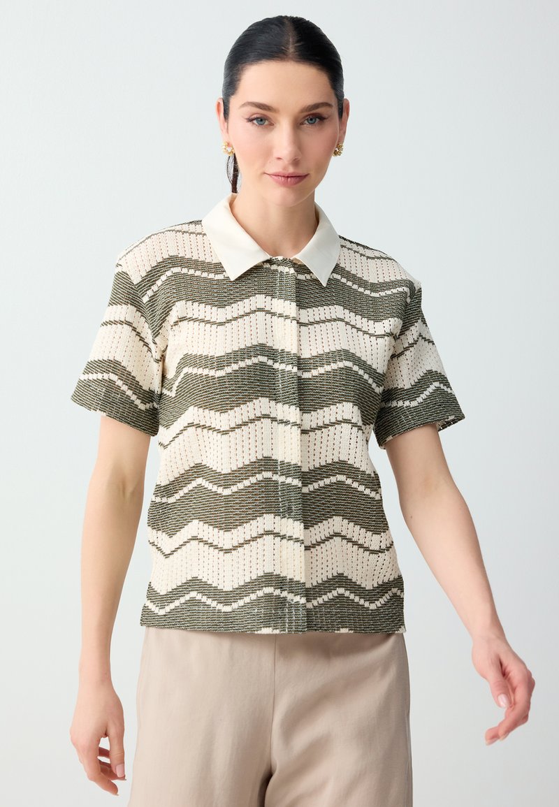 Femme portant un haut en maille à manches courtes avec col, rayé ondulé vert olive et crème, associé à un pantalon beige clair sur un fond uni.