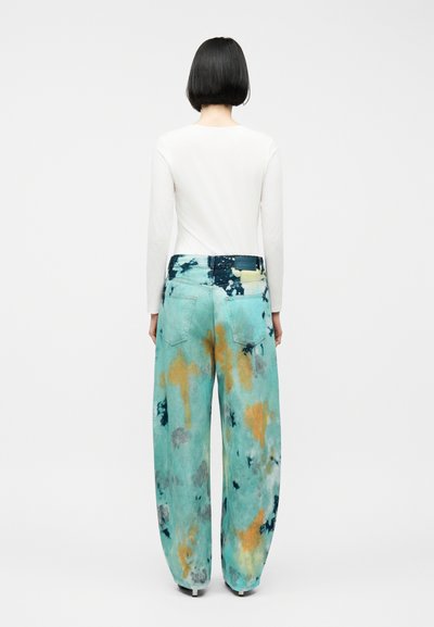 Γυναίκα με κοντά μαύρα μαλλιά, που φοράει λευκό στενό μακρυμάνικο τοπ και φαρδιά παντελόνια με τιρκουάζ γραβάτα-χρωματισμό με πορτοκαλί και γκρι κηλίδες, στέκεται με την πλάτη ορατή.