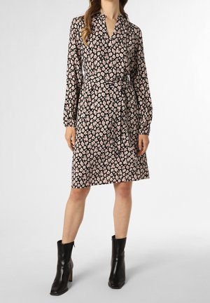 Vrouw draagt een knie-lange, lange mouwen jurk met een roze en beige bloemenpatroon op een zwarte achtergrond, gecombineerd met zwarte enkellaarsjes.