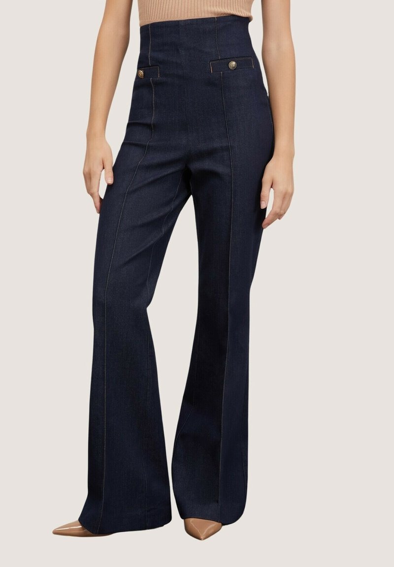 Femme portant un jean taille haute bleu foncé à jambes larges avec des détails de boutons près des poches et des chaussures beiges pointues.