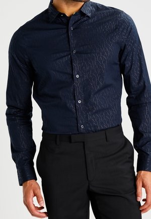 Mand iført en tætsiddende marineblå langærmet skjorte med subtil Armani-tekstprint, kombineret med sorte pæne bukser.