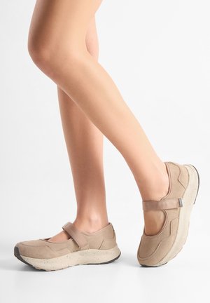 Nohy v béžových teniskách v štýle Mary Jane s hrubými bodkovanými podrážkami, jedna noha položená plochá a druhá mierne zdvihnutá na bielom pozadí.