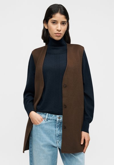 Καφέ αμάνικο γιλέκο πάνω από σκούρο μπλε ζιβάγκο, συνδυασμένο με ανοιχτό μπλε φαρδιά τζιν. Το πουλόβερ έχει ριγωτή υφή, το γιλέκο διαθέτει κουμπιά.