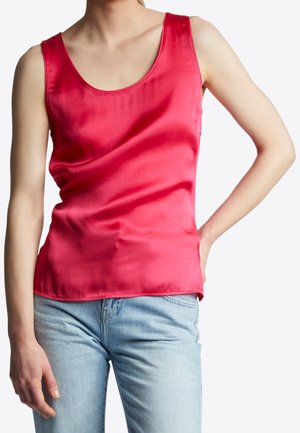 Kobieta ubrana w czerwoną, satynową, bezrękawnikową bluzkę i jasnoniebieskie dżinsy, stojąca z jedną ręką na biodrze na białym tle.