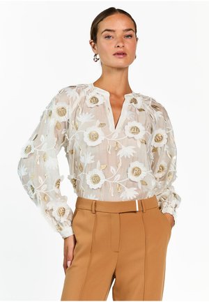 Vrouw die een doorschijnende witte blouse draagt met goudkleurige en witte bloemrijke borduursels, gecombineerd met hooggetailleerde camelkleurige broek en gouden oorringen.