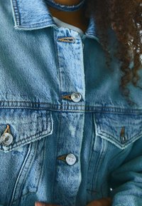 Primo piano di una persona che indossa una giacca di denim blu chiaro con bottoni d'argento, con i capelli ricci visibili sul lato destro.