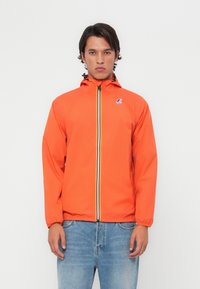 Chaqueta impermeable naranja con cierre de cremallera, puños elásticos y capucha con cordón. Presenta un detalle a rayas azules y amarillas.
