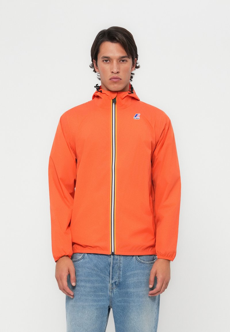 Chaqueta impermeable naranja con cierre de cremallera, puños elásticos y capucha con cordón. Presenta un detalle a rayas azules y amarillas.