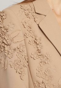 Blazer bej croit cu detalii florale aplicate texturate. Prezintă accente cusute și un guler decupat pentru un design structurat.