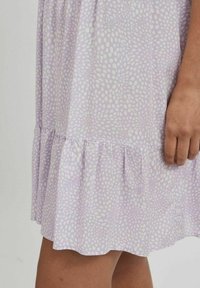 Levandulová sukně s bílým nepravidelným puntíkováním a volánkovým lemem, zachycena od pasu do půli stehna, s pravou rukou uvolněnou podél těla.