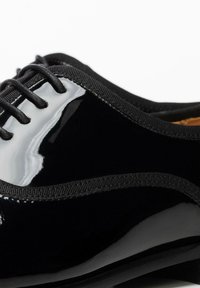 Chaussure Oxford en cuir verni noir avec lacets plats, accents mats texturés et finition brillante contrastante sur la tige.