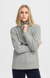 Suéter de punto texturizado en gris claro, con cuello alto y mangas largas. El modelo posa con los brazos cruzados, mostrando el corte holgado y los detalles del tejido.