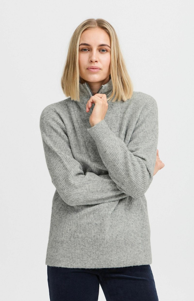 Suéter de punto texturizado en gris claro, con cuello alto y mangas largas. El modelo posa con los brazos cruzados, mostrando el corte holgado y los detalles del tejido.