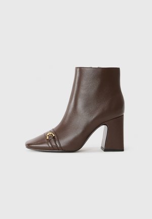 Bottines en cuir marron avec un bout pointu, accessoires en or et un talon épais. Texture lisse avec des détails de couture subtils.