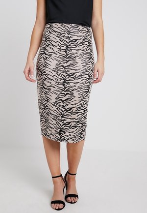 Kobieta w ołówkowej spódnicy w beżowo-czarny wzór tygrysa, nosząca czarne sandały na kostkę, stoi na tle jasnego, jednolitego tła.