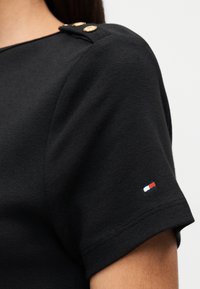 Μαύρο τοπ με κοντά μανίκια, δύο χρυσά κουμπιά στον ώμο και ένα μικρό λογότυπο σε κόκκινο, άσπρο και μπλε κοντά στο τελείωμα του μανικιού.