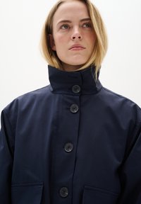 Laineen sininen vedenpitävä takki korkealla kauluksella, viisi mustaa nappia ja kaksi suurta etutaskua. Sileä tekstuuri ja minimalistinen muotoilu.