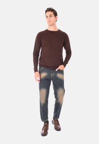 Maglione a maglia marrone abbinato a jeans blu sbiaditi con macchie più chiare, e scarpe marroni con superficie texture e suola nera.