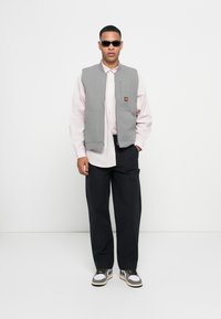 Γκρι αμάνικο γιλέκο πάνω από ένα ανοιχτό ροζ πουκάμισο με κουμπιά, σε συνδυασμό με σκούρα παντελόνια και αθλητικά παπούτσια, με λογότυπο στο γιλέκο και σκούρα γυαλιά ηλίου.