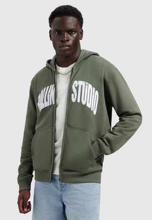 Jonge man met een olijfgroene zip-up hoodie met witte "ROLLING STUDIO" tekst, lichtblauwe spijkerbroek en een gouden ketting.