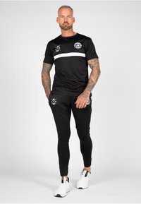 Black athletic t-shirt with white stripe and logo, paired with black fitted pants and white sneakers. Features tattoos and a neutral expression.