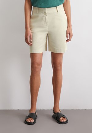 Shorts beige clair avec une coupe droite, des poches avant et un design plat à l'avant. Portés avec des sandales noires à glissière sur un sol neutre.