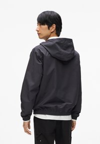 Fekete szélálló dzseki kapucnival, rugalmas mandzsettákkal és alsó szegéllyel. Simább textúra, könnyű anyag és laza szabás.