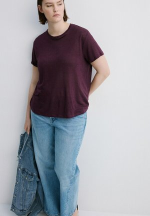 Frau trägt ein dunkelviolettes T-Shirt und hellblaue Jeans, hält eine Jeansjacke und steht mit neutralem Gesichtsausdruck vor einer weißen Wand.