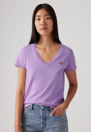 Jeune femme aux longs cheveux foncés portant un t-shirt lavande à col en V avec un petit logo rouge Levi’s et un jean bleu, debout devant un fond uni.