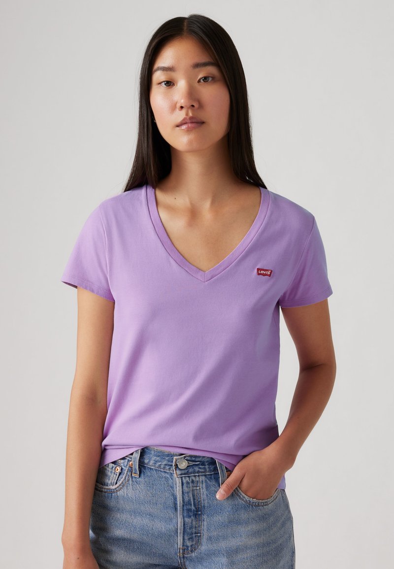 Jeune femme aux longs cheveux foncés portant un t-shirt lavande à col en V avec un petit logo rouge Levi’s et un jean bleu, debout devant un fond uni.