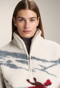 Fleecová mikina s vysokým golierom, krémovým základom, modrými pruhmi a vyšívaným červeným motívom. Má biely zips a mäkkú, textúrovanú tkaninu.