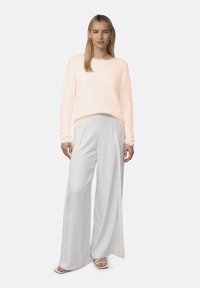 Lichtroze trui met lange mouwen, gecombineerd met lichtgrijze broek met wijde pijpen en witte sandalen, tegen een neutrale achtergrond.