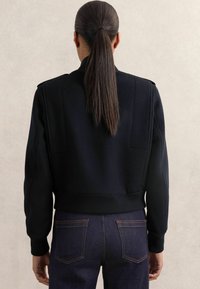 Personne avec de longs cheveux noirs attachés en queue de cheval, portant une veste noire et un jean bleu foncé, se tenant contre un fond clair et uni.