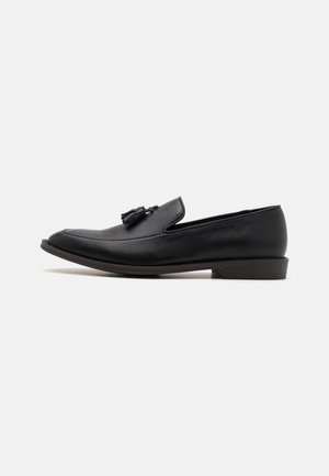 Black leather loafers with a rounded toe and flat sole, featuring a decorative tassel on the vamp and minimal stitching detail.