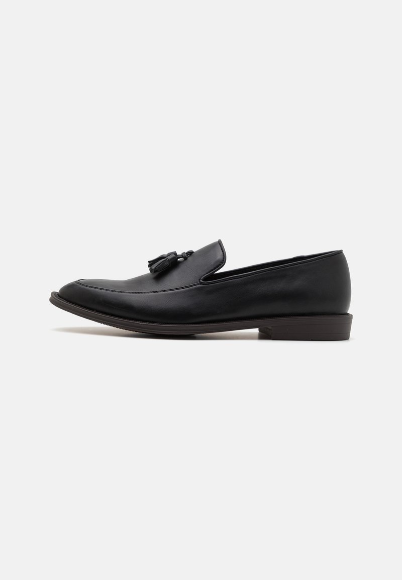Mocassins en cuir noir avec un bout rond et une semelle plate, présentant un gland décoratif sur la tige et un minimum de détails de couture.