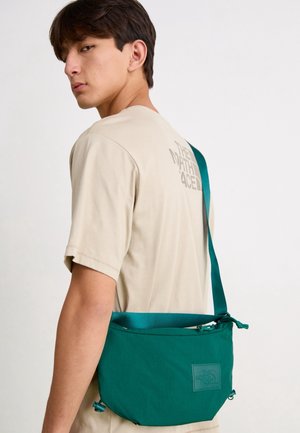 Jeune homme aux cheveux bruns courts portant un t-shirt beige The North Face et portant un sac bandoulière turquoise avec logo sur le devant, sur fond blanc.