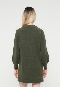 Kvinne med langt blondt hår som har på seg en olivengrønn strikket langermet kjole, stående mot en enkel hvit bakgrunn, med ryggen mot betrakteren.
