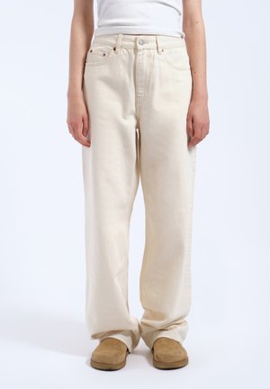 Person iført løstsiddende beige bukser, hvid top og beige slip-on sko står mod en hvid baggrund.