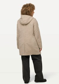 Cappotto in lana beige con cappuccio, caratterizzato da una vestibilità dritta e uno spacco nella parte posteriore. Abbinato a pantaloni larghi scuri e scarpe nere.