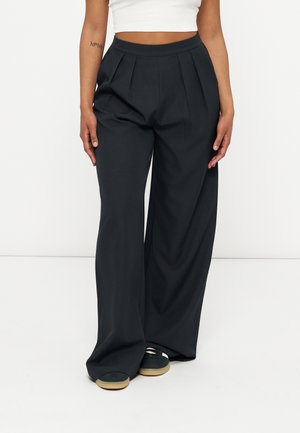 Personne portant un pantalon noir à jambes larges avec des plis et des baskets noires à semelles beiges, associée à un haut court blanc.